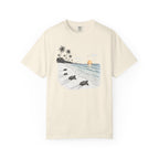 An off-white short-sleeve crewneck t-shirt featuring a central graphic print of a tropical beach scene with palm trees, sea turtles on the sand, and a sunset over the ocean.