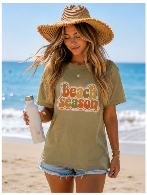 Woman on a beach wearing a 'Beach Season' t-shirt with a straw hat and water bottle.