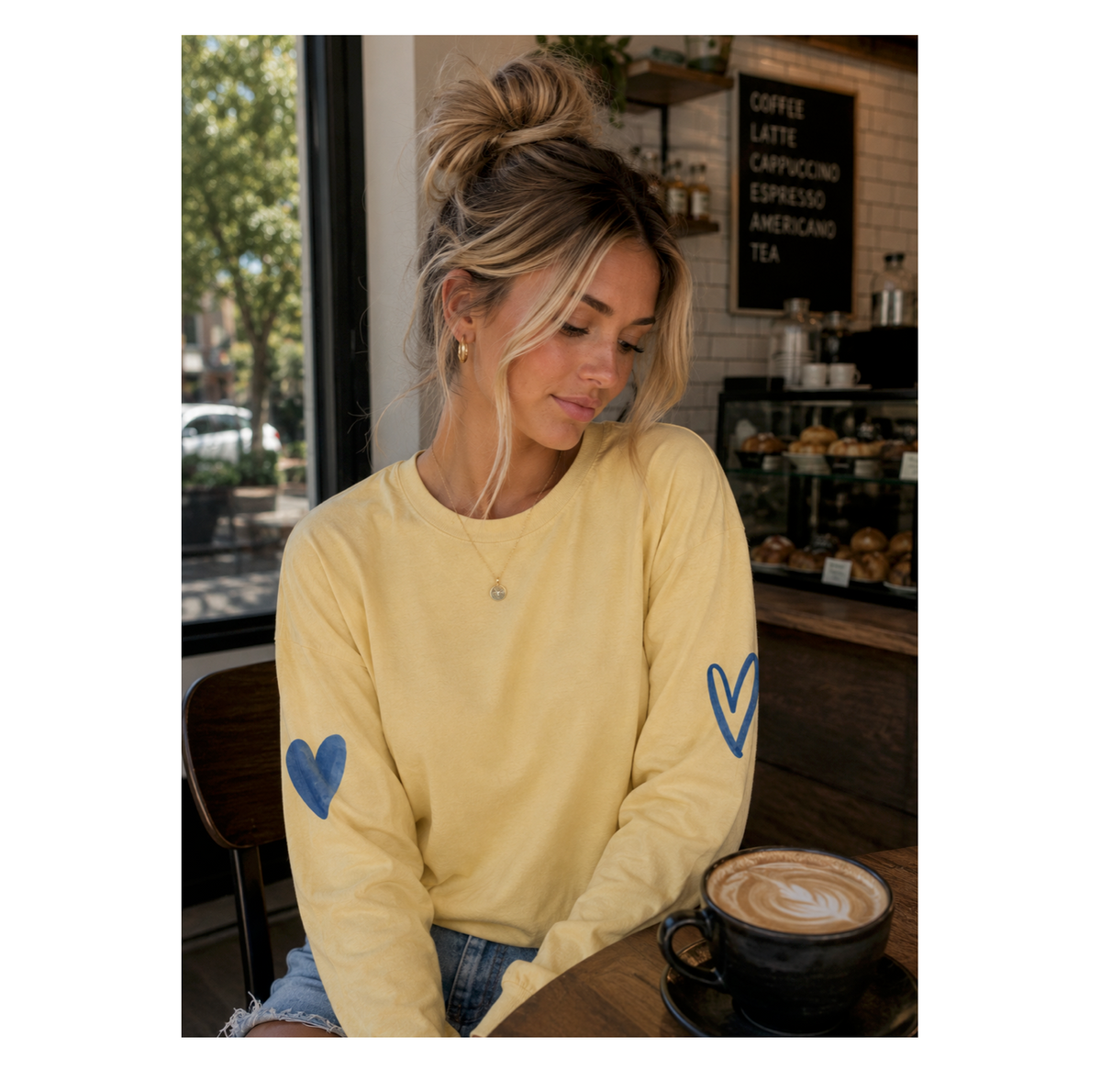 A woman sitting at a cafe table wearing a pale yellow long-sleeve shirt with blue heart designs on both sleeves, paired with blue jeans and a black coffee cup in front of her.