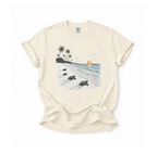 An off-white short-sleeve t-shirt with a graphic print of a tropical beach scene featuring sea turtles, palm trees, and a sunset, laid flat on a white background.