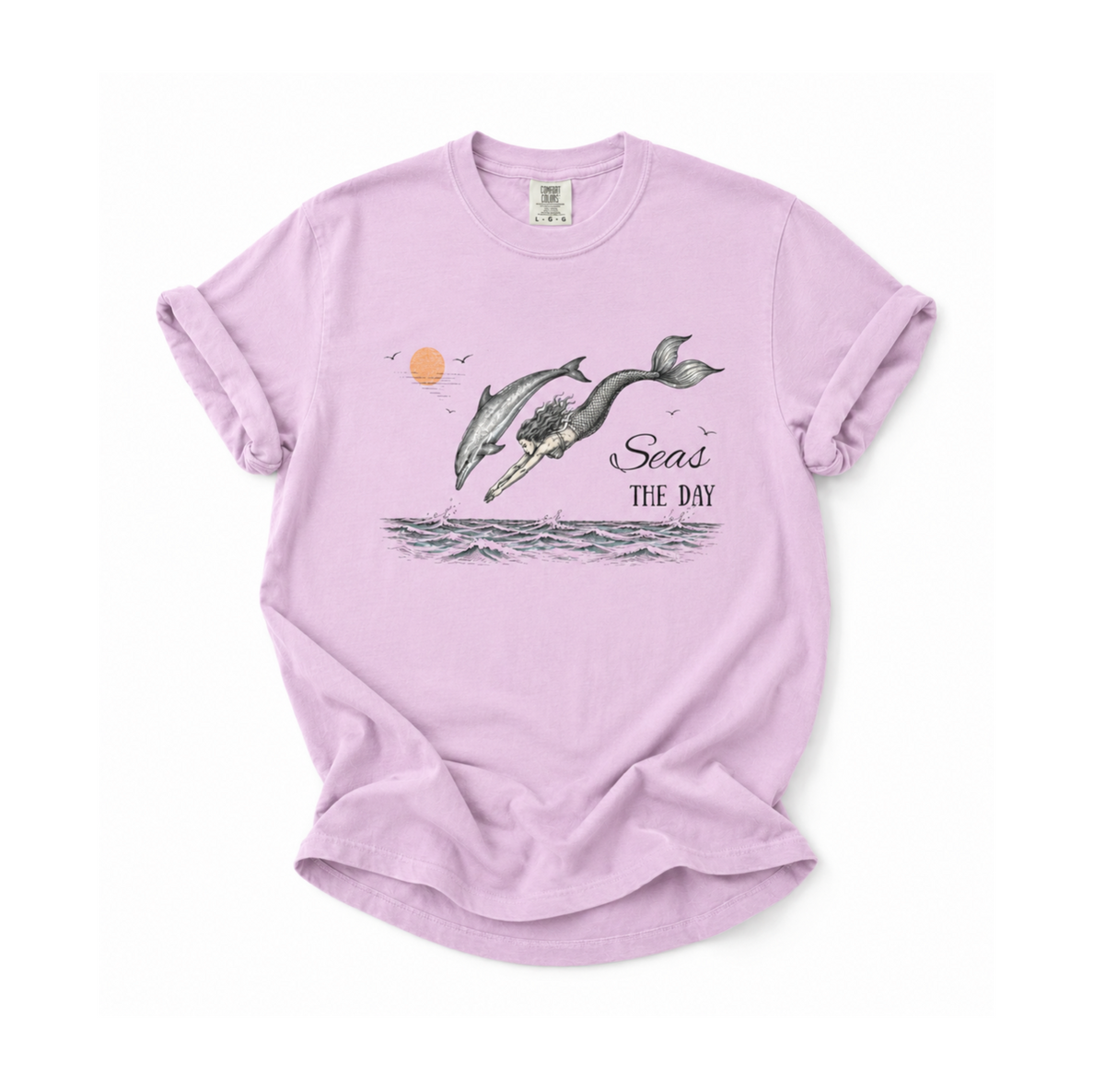 A light purple short-sleeve t-shirt with a graphic of two whales jumping out of the water, a small orange sun, and the text 'Seas THE DAY'.