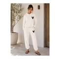 A woman modeling a matching cream-colored lounge set consisting of a pullover hoodie and jogger pants, both featuring a black sequined heart on the chest and thigh. She is standing in a sunlit outdoor patio setting next to a potted olive tree.