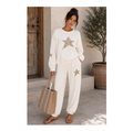 A woman stands outdoors wearing a matching cream lounge set with metallic gold star graphics on the sweatshirt and pants. She is holding a large beige tote bag and wearing brown sandals, set against a rustic background with a wooden door and a potted tree.