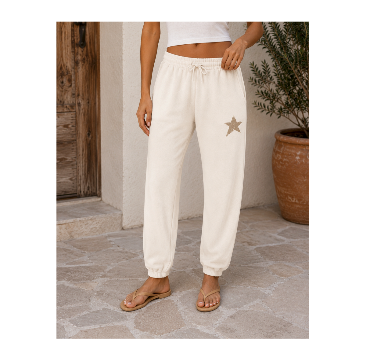 A person wearing cream-colored jogger sweatpants with a drawstring waist and a small star patch on the left thigh, standing on a stone patio next to a potted plant.