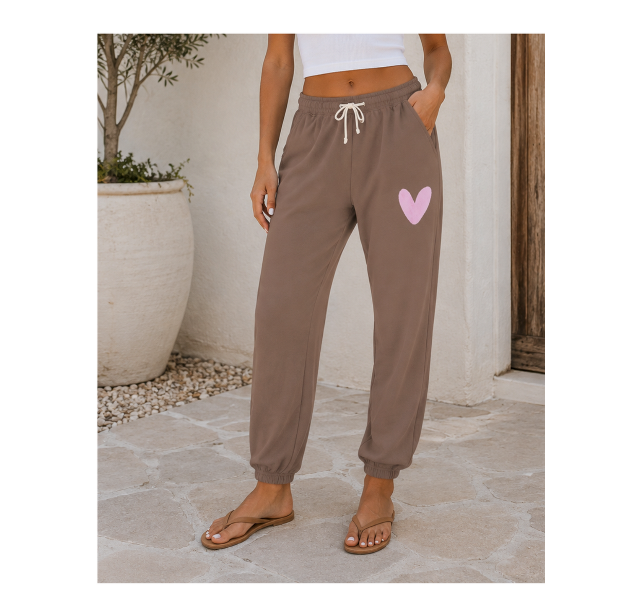 A person wearing brown jogger-style sweatpants with a white drawstring and a light pink heart graphic on the upper thigh, standing outdoors on a stone patio.